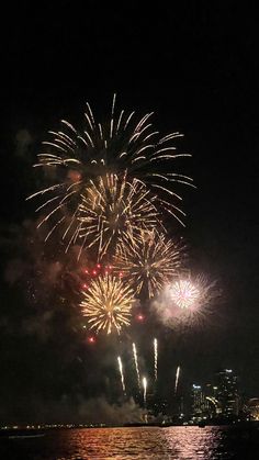 fireworks are lit up in the night sky over water and city buildings on the other side