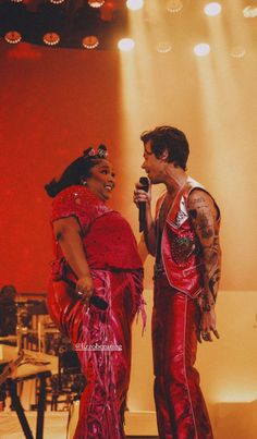 two people standing on stage with microphones in their hands and one person wearing red