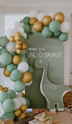 a table topped with balloons next to a green dinosaur balloon arch filled with gold and white balloons