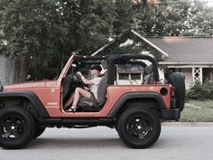 a man and woman sitting in the driver's seat of a red jeep parked next to a house