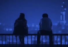 two people are sitting on a railing looking at the city lights in the night sky