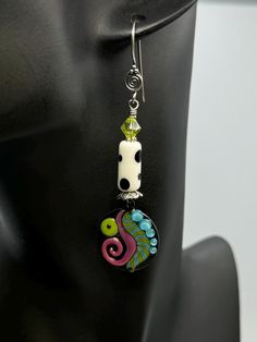 These earrings feature a unique and artistic design. It begins with a sterling silver ear hook that leads down to a small, intricately crafted silver spiral. Below this, a bright lime-green Swarovski faceted crystal adds a pop of color and sparkle.  The next component is a white cylindrical lampwork bead with bold black polka dots, giving the piece a playful and modern feel.  The earring's focal point is a round, black enameled copper pendant adorned with vibrant, abstract patterns: a swirling pink shape, a lime-green dot, and blue bubble-like accents alongside wavy green and blue lines, creating an eye-catching and whimsical design.  The combination of colors and shapes makes this earring a statement piece that blends contemporary and artistic styles. Earrings measure 3" long Will come gi Lampwork Bead Earrings, Playful Jewelry, Enameled Copper, Copper Pendant, Colorful Earrings, Copper Pendants, Ear Hook, Faceted Crystal, Bold Black