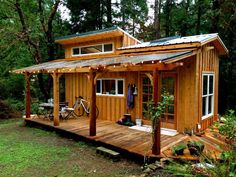 a small wooden cabin in the woods with a porch and covered area for bikes to sit on