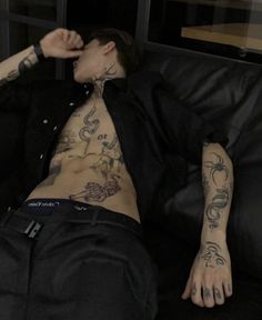 a man with tattoos on his chest laying on a black leather couch in front of a window