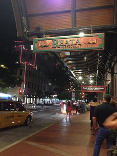 people are walking down the sidewalk under an awning at night time, with cars parked on the street below