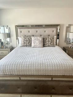 a white bed with pillows on it and two mirrors in the corner behind it, along with other furniture