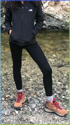 a woman in black jacket and red shoes standing on rocks next to water with her legs crossed