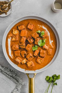 a white bowl filled with curry and tofu on top of a table next to some herbs