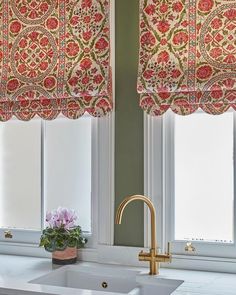 a kitchen sink sitting under two windows next to a window covered in red and green curtains