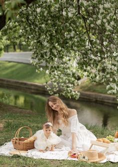 a woman and her child sitting on a blanket in the grass near a river with picnic food