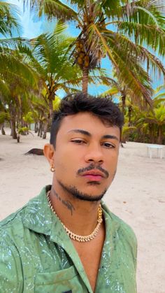 a man wearing a green shirt and gold necklace standing in front of palm trees on the beach