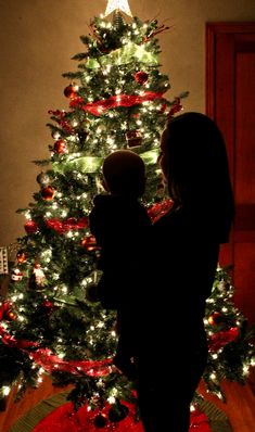 a mother and her child standing in front of a christmas tree with lights on it