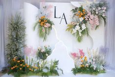 a white backdrop with flowers and greenery in front of the letters la on it