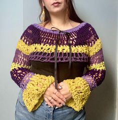 a woman is standing in front of a wall with her hands on her hips wearing a purple and yellow crochet sweater