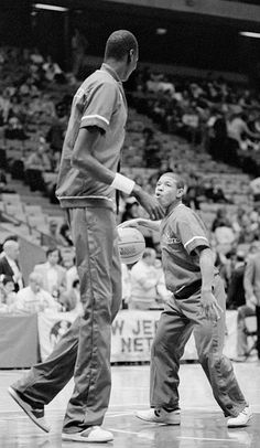 two men are playing basketball in front of an arena full of people, one holding a ball and the other looking up