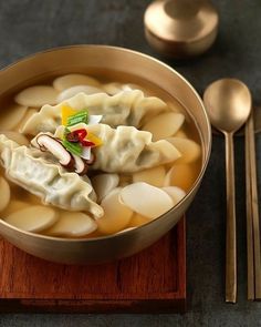 a bowl of soup with dumplings and spoons next to it on a wooden board