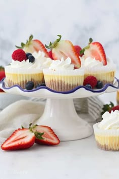 three cupcakes topped with whipped cream and strawberries on a white cake stand