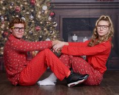 two people sitting on the floor in pajamas near a christmas tree with presents behind them