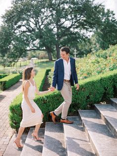 a man and woman are walking down some steps in front of the bushes, holding hands