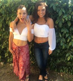 two young women standing next to each other in front of some bushes and green plants