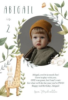 a baby is wearing a hat and standing next to a giraffe with leaves around it