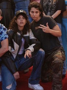 two young men sitting next to each other on a red carpet with people standing behind them