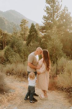 a man and woman holding a small child while standing on a dirt road in the woods