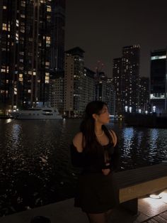 a woman standing next to a body of water in front of tall buildings at night