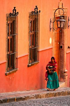 a woman walking down the street in front of an orange building with two windows on each side