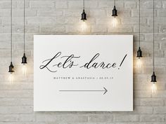 a welcome sign hanging from the side of a white brick wall with lights around it