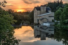 the sun is setting over an old factory building on the water's edge with trees around it