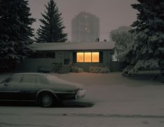 a car parked in front of a house on a snow covered street with trees and buildings behind it