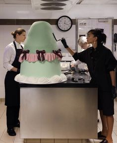 two women standing in front of a giant cake on a counter top with the woman decorating it