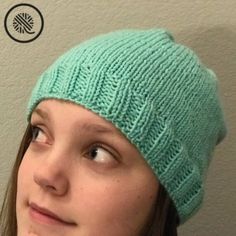 a woman wearing a green knitted beanie looks up at the camera while standing in front of a wall