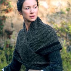 a woman wearing a knitted shawl in front of a stone wall and bushes