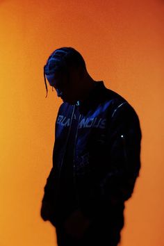 a man with dreadlocks standing in front of an orange wall wearing a black jacket