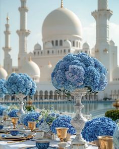 a table set with blue flowers and gold place settings in front of a large white building