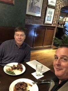 two men sitting at a table with plates of food in front of them, smiling for the camera