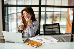 a woman sitting at a table talking on her cell phone and holding a cup of coffee