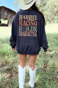 [Image description: A person is standing in a grassy field, facing away from the camera, wearing a dark sweatshirt that reads "BARREL RACING BUILDS CHARACTER" in large, rustic-style text. They are also wearing a cowboy hat and tall white boots. A weathered barn is visible in the background, surrounded by trees under a bright blue sky.] Racing Horse, Rodeo Girls, Barrel Racer, Girls Jumpers, Cozy Gift, Barrel Racing