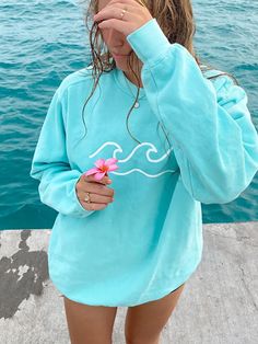 a woman standing next to the water holding a pink flower in her hand and wearing a blue sweatshirt