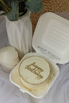 a birthday cake in a plastic container next to a vase with flowers on the table