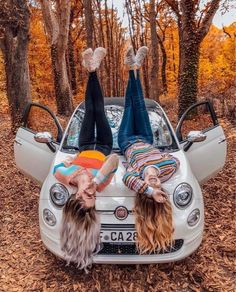 two women laying on the hood of a car in front of some trees and leaves