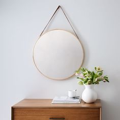 a white vase with flowers on a table next to a round mirror hanging on the wall