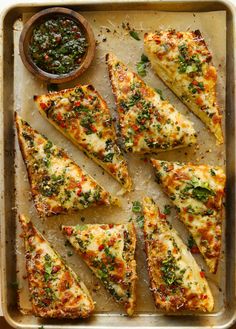 several slices of pizza on a baking sheet with sauce and herbs in a bowl next to it