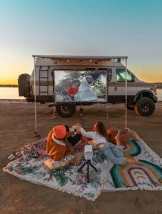 two people sitting on a blanket in front of a truck with a large tv screen