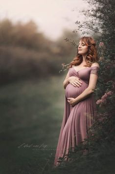 a pregnant woman in a pink dress standing next to a bush with her hands on her stomach