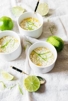 three white bowls filled with rice and limes on top of a cloth covered table