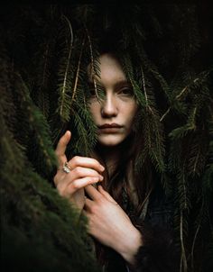 a woman with long hair is hiding behind some branches and looking at the camera while wearing a ring