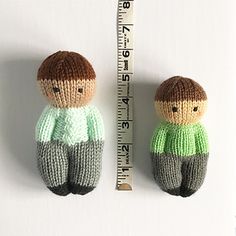 two knitted dolls are next to a measuring tape on a white surface, one is green and the other is brown